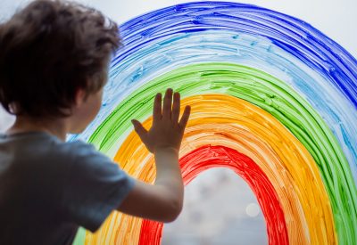 Image shows a child standing at a window, which has a rainbow painted on it.