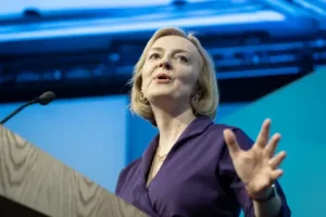 Liz Truss, a woman with a blonde bob in a purple shirt, stands against a blue background at a lecturn