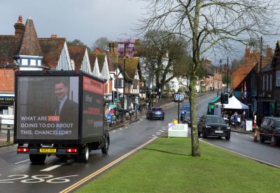 An ad van drives past a grassy area in a town, with an image of Jeremy Hunt under the words "what are you going to do about it Chancellor?" on the other side it are words including "struggling, worried, concerned, expensive"