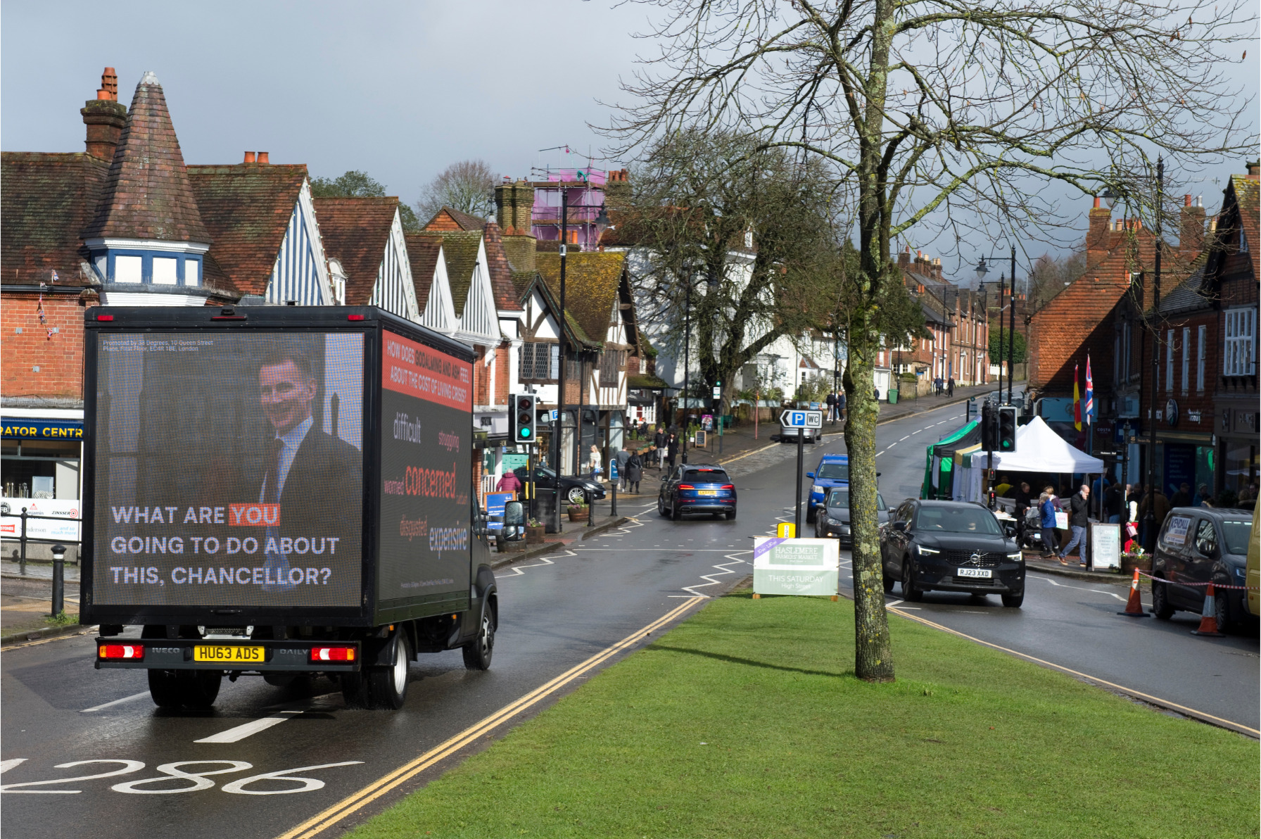 An ad van drives past a grassy area in a town, with an image of Jeremy Hunt under the words "what are you going to do about it Chancellor?" on the other side it are words including "struggling, worried, concerned, expensive"