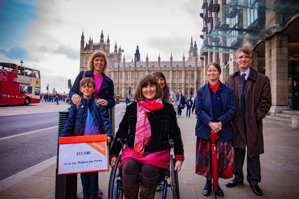 A group of people stand in front of the Houses of Parliament. One of them, a young boy, holds a petition box saying "Save our Parks".