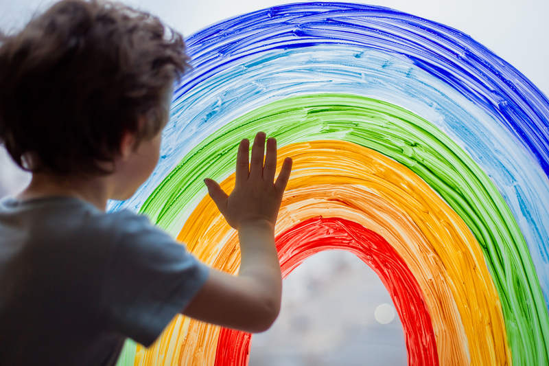 Image shows a child standing at a window, which has a rainbow painted on it.