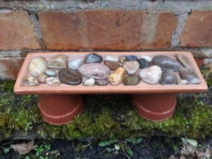 A shallow dish filled with clean stones is placed on top of two upturned plant pots. There is water in the dish.