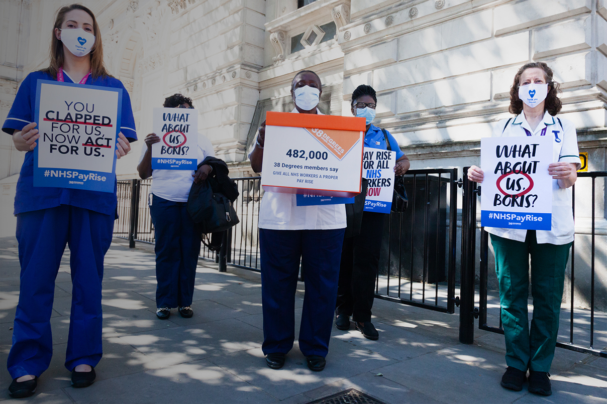 A photo of a group of NHS workers outside Downing Street, about to hand in a petition calling for NHS workers to receive a proper pay rise. They are holding posters with campaign messages and are socially distanced from one another and wearing face masks.