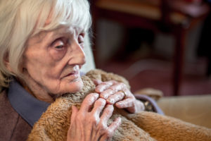 Photo of an older woman with white hair clutching a blanket towards her, with a despondent expression.