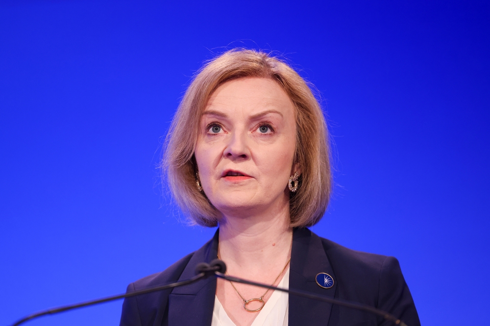 Liz Truss, a woman with a blonde bob in a black jacket, stands against a blue background giving a speech