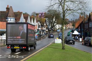An ad van drives past a grassy area in a town, with an image of Jeremy Hunt under the words "what are you going to do about it Chancellor?" on the other side it are words including "struggling, worried, concerned, expensive"