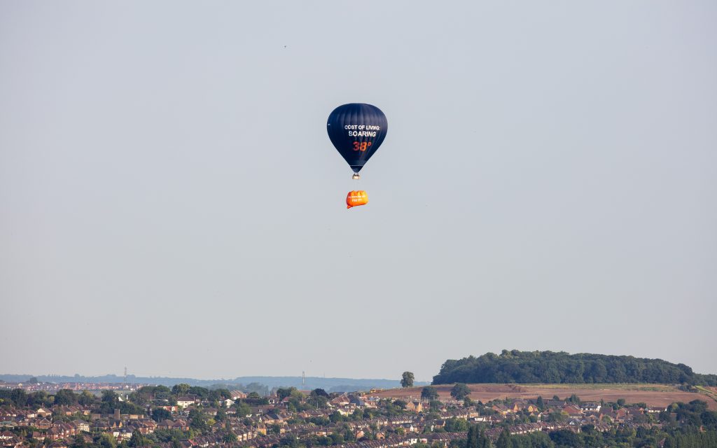 A blue hot air balloon with the phrase "the cost of living: soaring" flying over a city